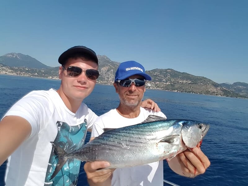 Deux hommes souriants tenant un gros poisson argenté sur un bateau avec la côte en arrière-plan.