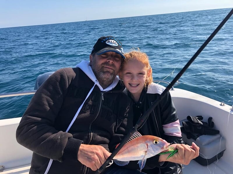 Homme et jeune fille tenant un poisson rose sur un bateau, tous deux souriants en mer.