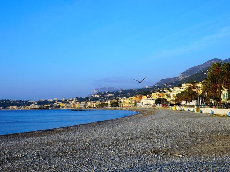 Plage de galets avec mer calme, ville et montagnes en arrière-plan sous un ciel bleu clair.