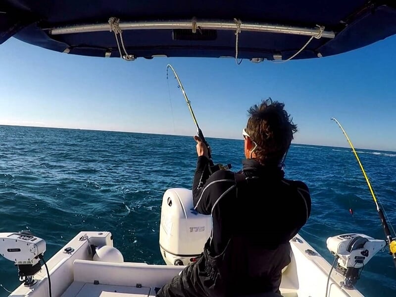 Pêcheur en mer tenant une canne à pêche pliée, tentant de remonter un poisson dans un bateau.