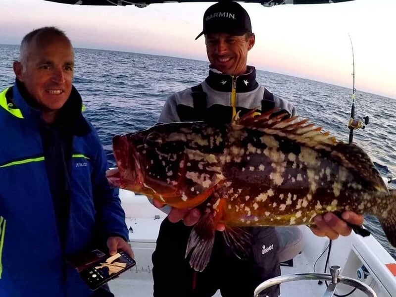 Deux hommes souriants à bord d'un bateau, l'un tenant un gros poisson coloré fraîchement pêché.