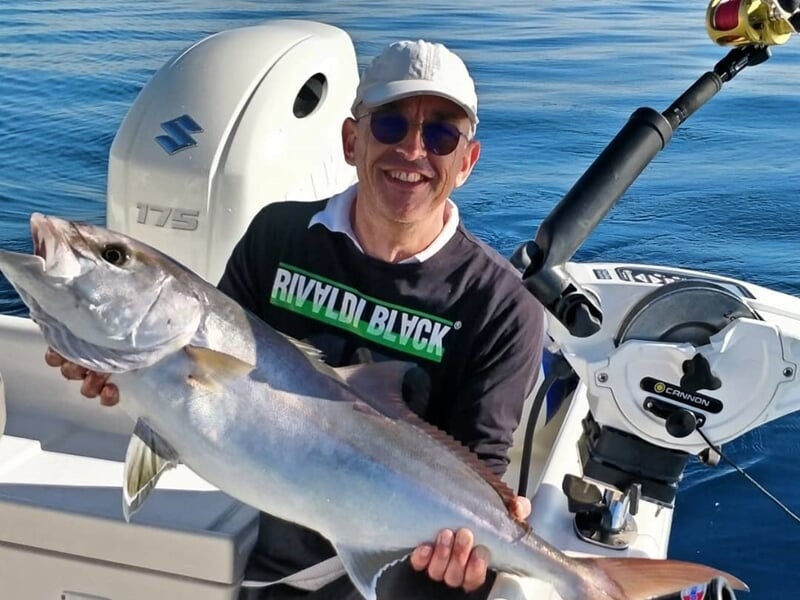 Homme souriant tenant un gros poisson argenté à bord d'un bateau de pêche en mer calme.