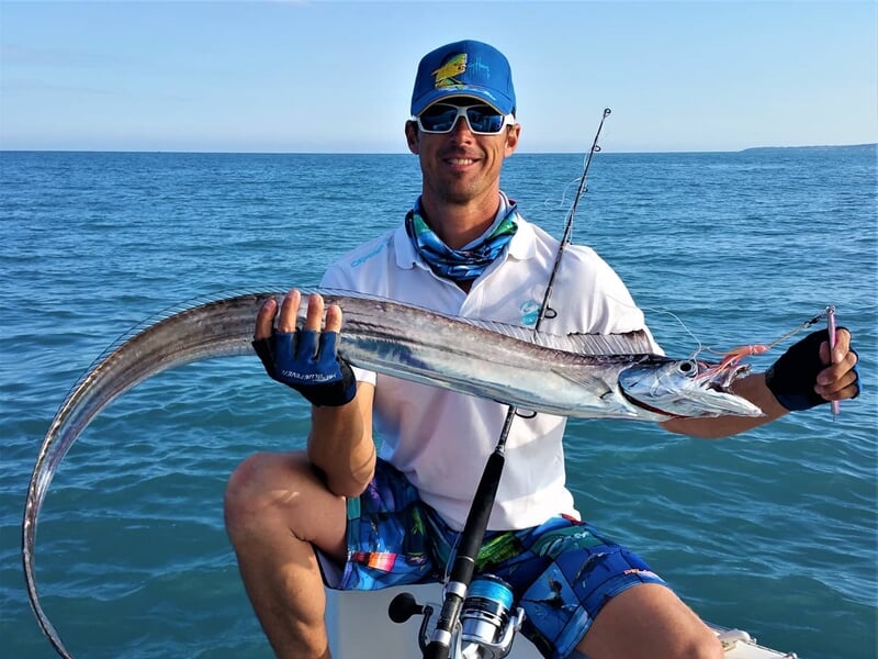 Pêcheur assis sur un bateau montrant un long poisson argenté avec une bouche ouverte et dents visibles.