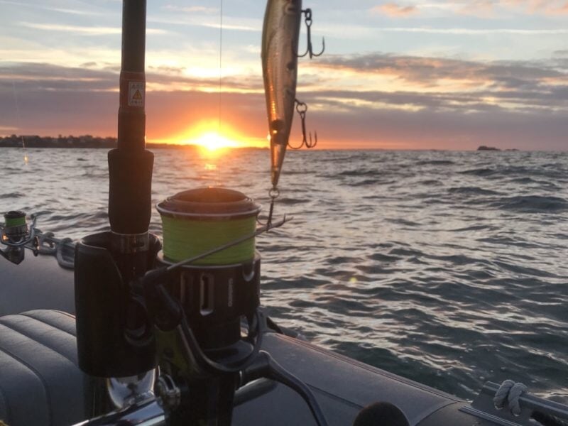 Une canne à pêche équipée d’un leurre suspendu est posée sur le bord d’un bateau, capturant le moment précis du coucher de soleil à l’horizon, avec des reflets dorés sur une mer légèrement agitée.