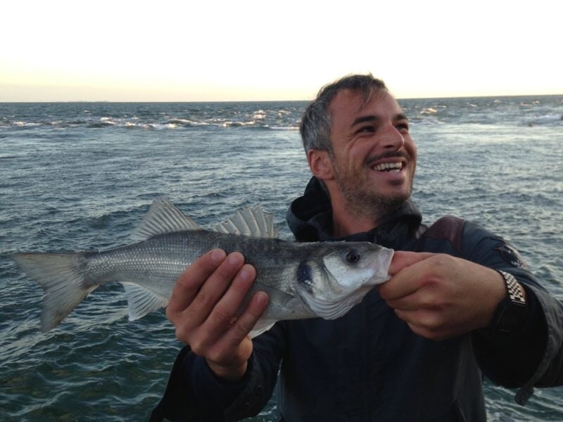 Un homme souriant, debout sur les rochers en bord de mer, tient un bar pêché à la main, les vagues se brisant derrière lui sous un ciel de fin de journée.