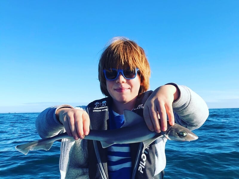 Un enfant aux cheveux roux et portant des lunettes de soleil bleues tient un petit requin par les nageoires, en souriant sur un bateau au large, avec une mer calme et un ciel d’un bleu intense.