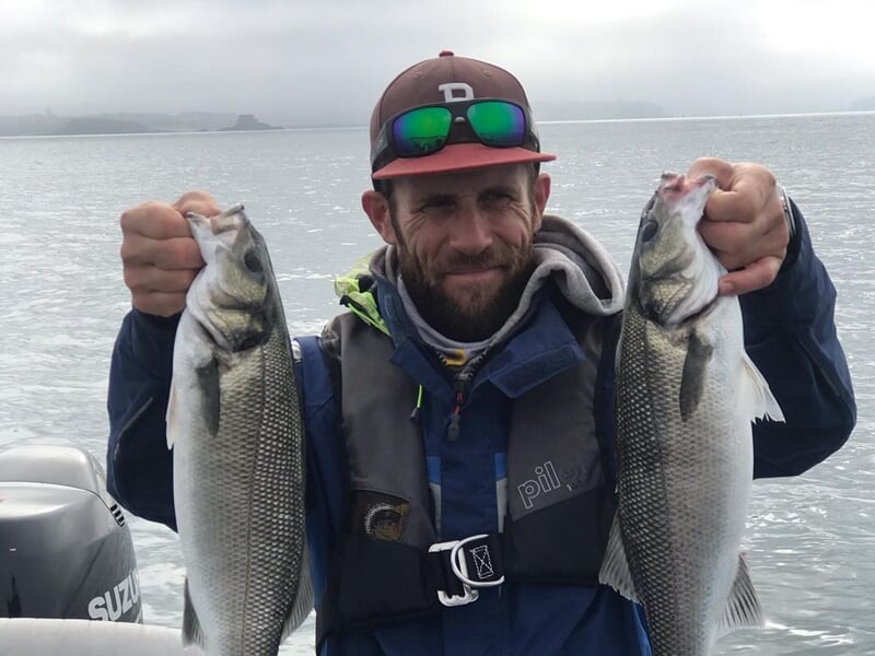Un pêcheur souriant, équipé d’un gilet de sauvetage et d’une casquette, présente deux bars de taille moyenne, un dans chaque main, sur un bateau à moteur en mer avec un ciel couvert à l’arrière-plan.