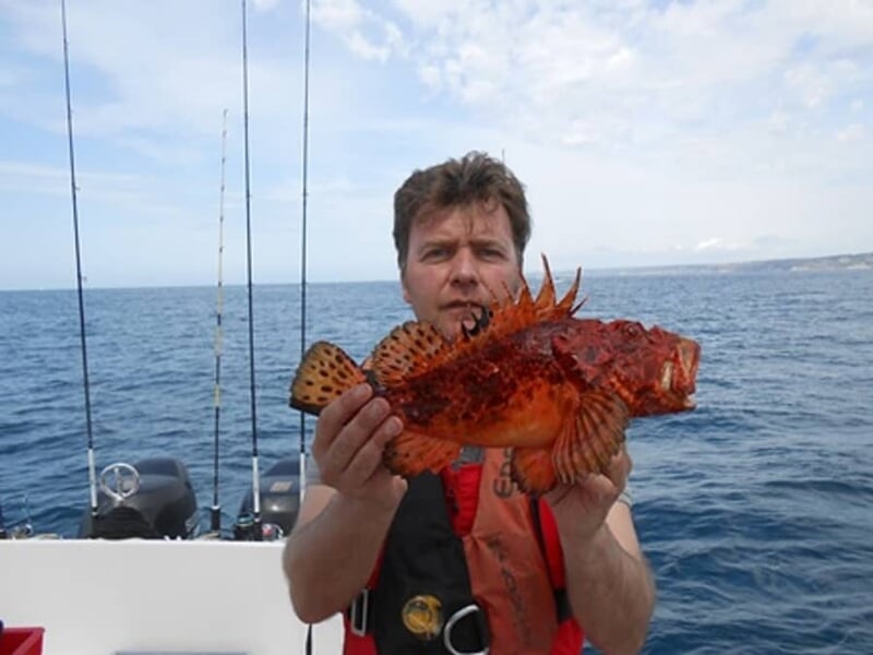 Un homme en gilet de sauvetage tient un gros poisson rouge épineux sur un bateau en mer, avec cannes à pêche en arrière-plan.