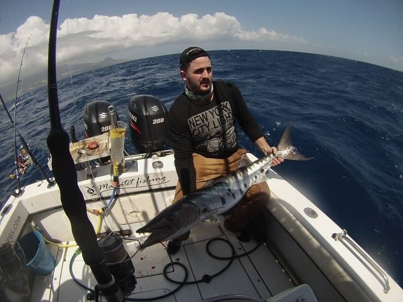 Homme assis dans un bateau tenant un gros barracuda argenté, moteur hors-bord et mer en arrière-plan.