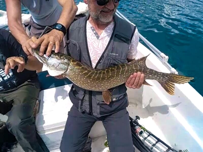 Homme tenant un grand poisson sur un bateau, entouré de deux autres personnes, sur un lac aux eaux calmes.