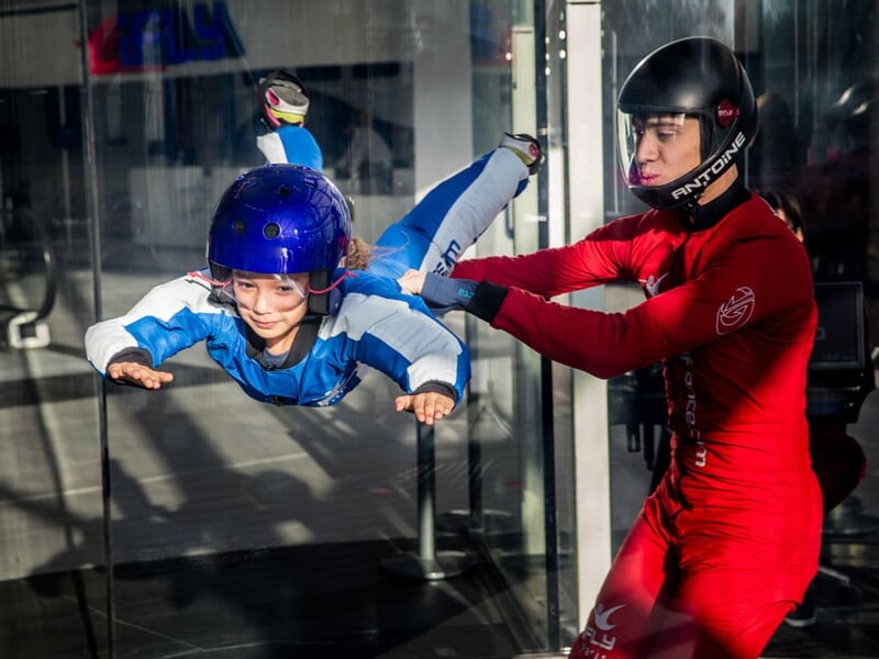 Enfant en combinaison bleue et casque violet en chute libre simulée dans un tunnel à vent avec un instructeur en rouge.