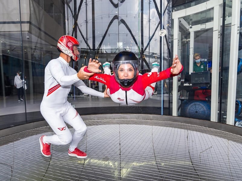 Femme en combinaison blanche et rouge en chute libre simulée dans un tunnel à vent avec un instructeur en blanc.