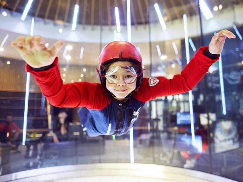 Jeune participant en combinaison de vol rouge et casque, volant avec confiance dans un simulateur de chute libre, les lumières se reflétant sur le tube en verre