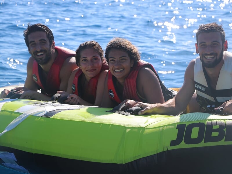Quatre personnes souriantes sur une bouée tractée verte sur la mer à Nice, portant des gilets de sauvetage.