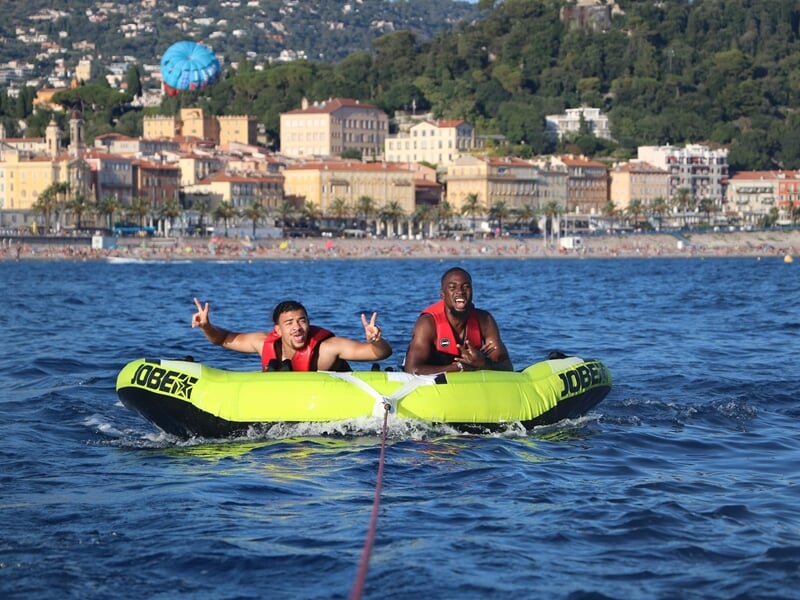 Deux hommes sur une bouée tractée verte sur la mer, faisant des signes de paix avec la ville de Nice en arrière-plan.