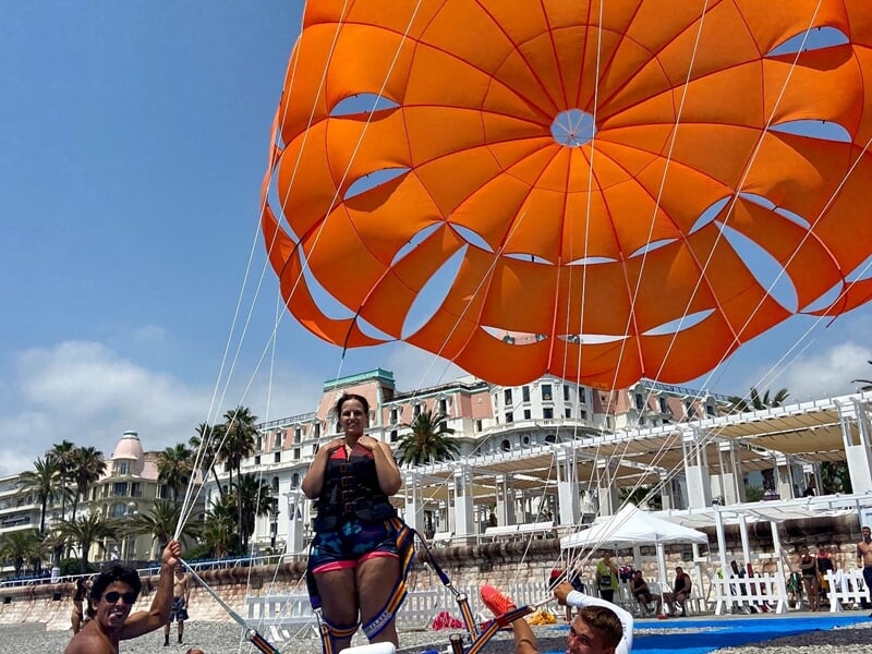 Personne en parachute ascensionnel orange au-dessus de la mer, avec un bateau et un hôtel en arrière-plan à Nice.