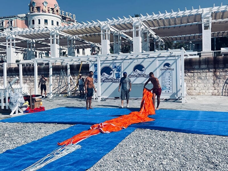 Préparation d'un parachute ascensionnel orange sur une plage avec plusieurs personnes autour, à Nice.