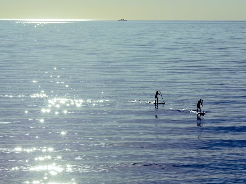 Deux personnes pratiquant le stand-up paddle sur une mer calme et scintillante sous un ciel clair.