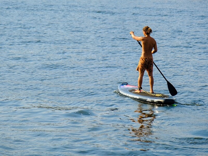 Homme debout sur une planche de stand-up paddle pagayant sur une mer calme avec un ciel clair.