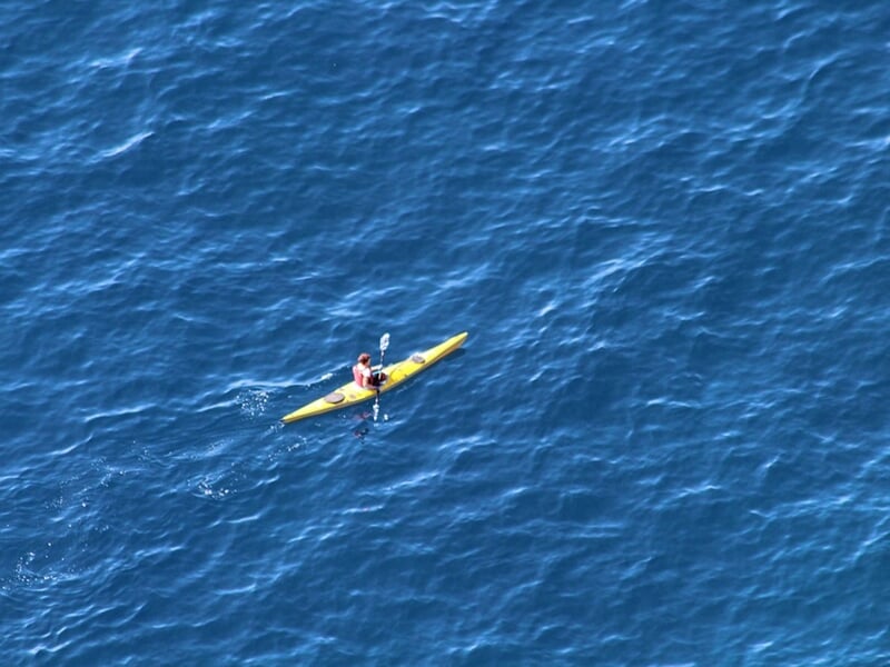 Personne pagayant seule en kayak sur une mer bleue, vue aérienne avec un paysage côtier en arrière-plan.