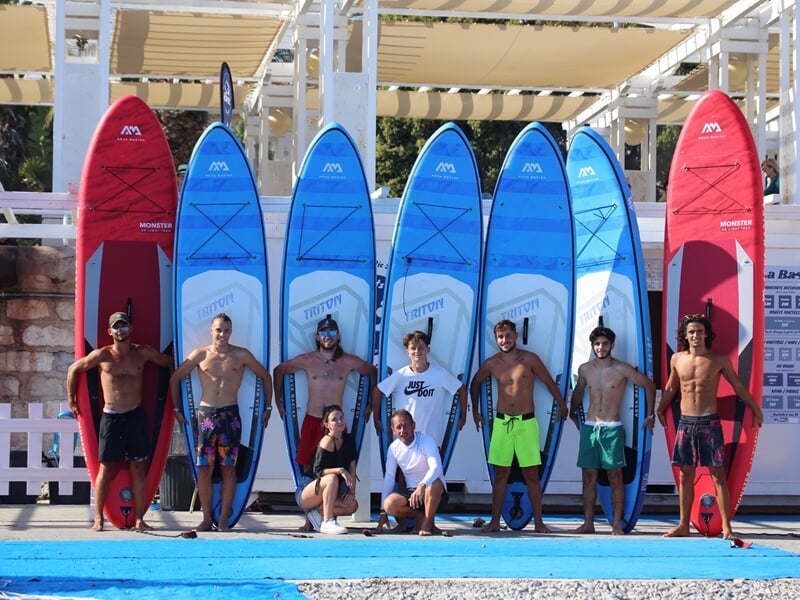 Groupe de personnes posant devant plusieurs planches de stand-up paddle colorées sur une plage à Nice.