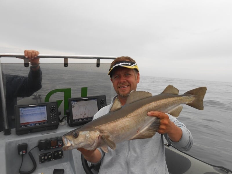 Homme souriant tenant un gros poisson pêché en mer sur un bateau avec équipements de navigation visibles.