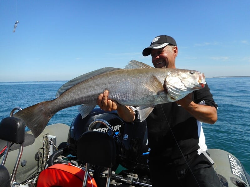 Homme debout sur un bateau tenant un poisson de grande taille pêché en mer sous un ciel bleu clair.