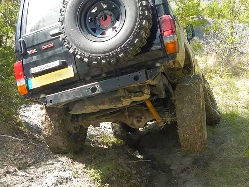 4x4 noir très incliné en montée boueuse, roue arrière gauche en l’air, roue de secours fixée à l’arrière