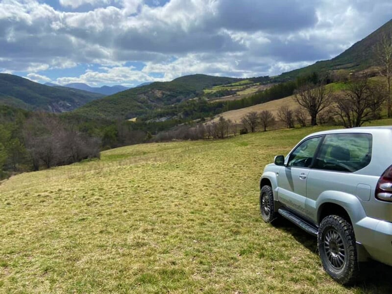 SUV gris stationné sur une prairie verdoyante entourée de montagnes et vallées sous ciel nuageux