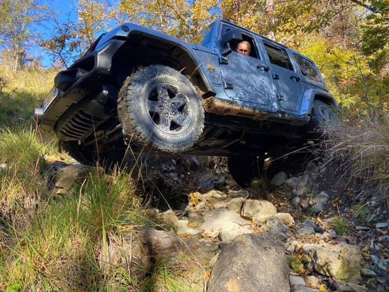 Jeep gris franchissant un passage rocheux en forêt, roue avant gauche en l’air, conducteur visible à la fenêtre