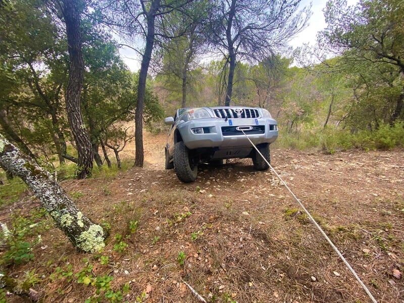 SUV gris treuillé à l’avant sur chemin forestier en pente entre arbres et broussailles