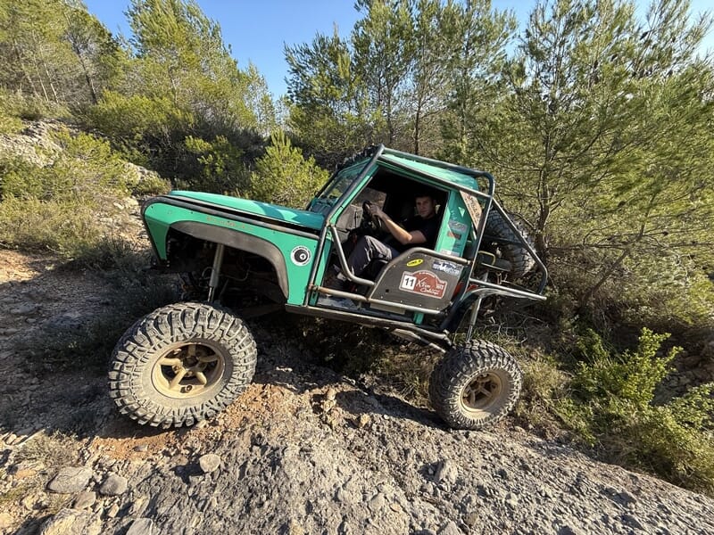 4x4 vert tout-terrain franchissant des rochers en pente dans un paysage boisé sous ciel bleu