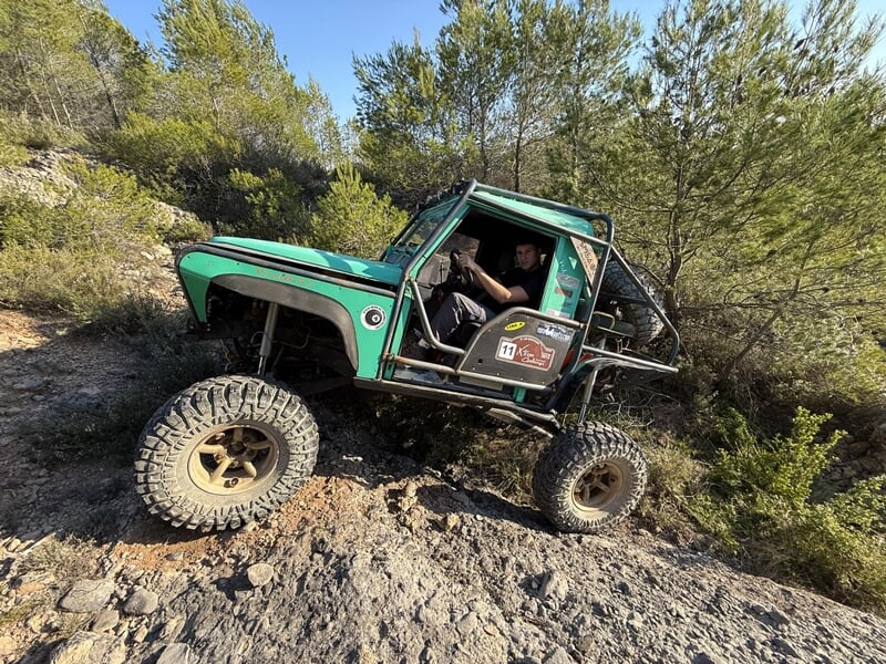 4x4 vert tout-terrain franchissant des rochers en pente dans un paysage boisé sous ciel bleu