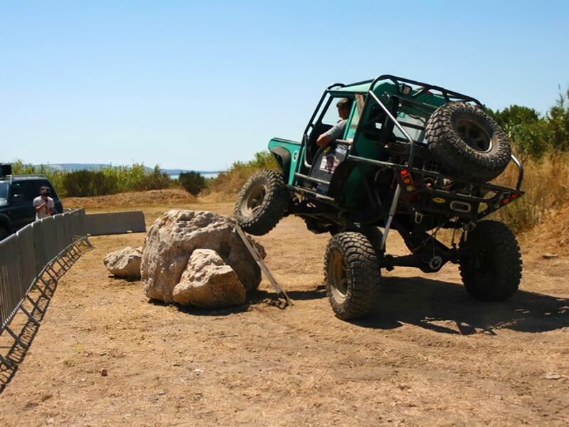 4x4 vert franchissant un obstacle rocheux sur un terrain sec avec des spectateurs derrière une barrière.
