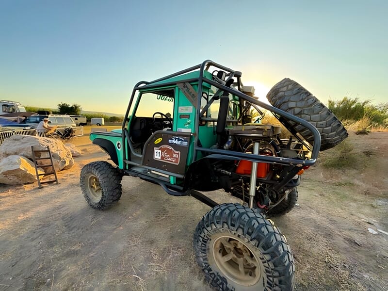 Vue arrière d'un 4x4 vert avec roue de secours, garé sur un terrain sec au coucher du soleil.