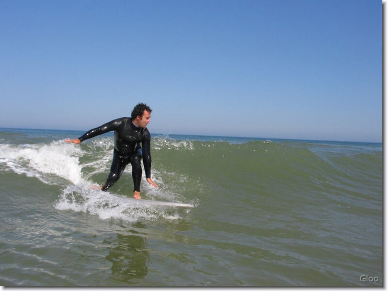 Homme en combinaison de surf noir chevauchant une petite vague sur une mer calme sous un ciel clair.