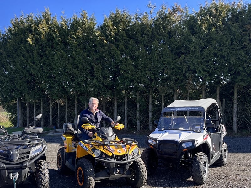 Homme souriant assis sur un quad jaune, entouré d'autres quads stationnés devant une haie de conifères.