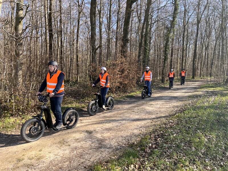 Groupe de personnes en gilets orange roulant en trottinettes électriques sur un chemin forestier au Mont Saint Adrien.