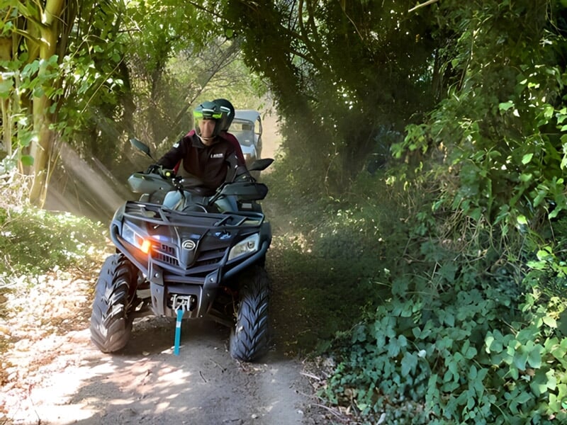 Personne conduisant un quad dans un sentier étroit entouré de végétation dense et de rayons de soleil.