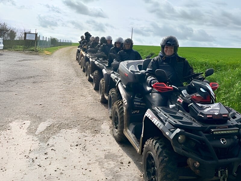 Groupe de personnes en quads alignés sur un chemin de campagne, avec un ciel bleu et des champs en arrière-plan.