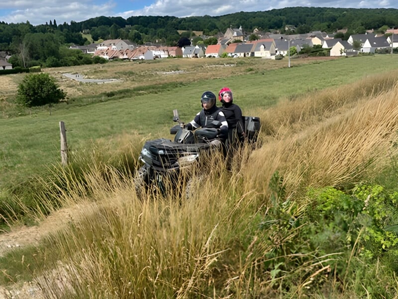 Deux personnes en quad roulant sur un chemin herbeux avec un village en arrière-plan sous un ciel clair.