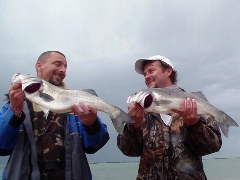 Deux hommes souriants tenant chacun un poisson fraîchement pêché au bord de la mer sous un ciel nuageux.