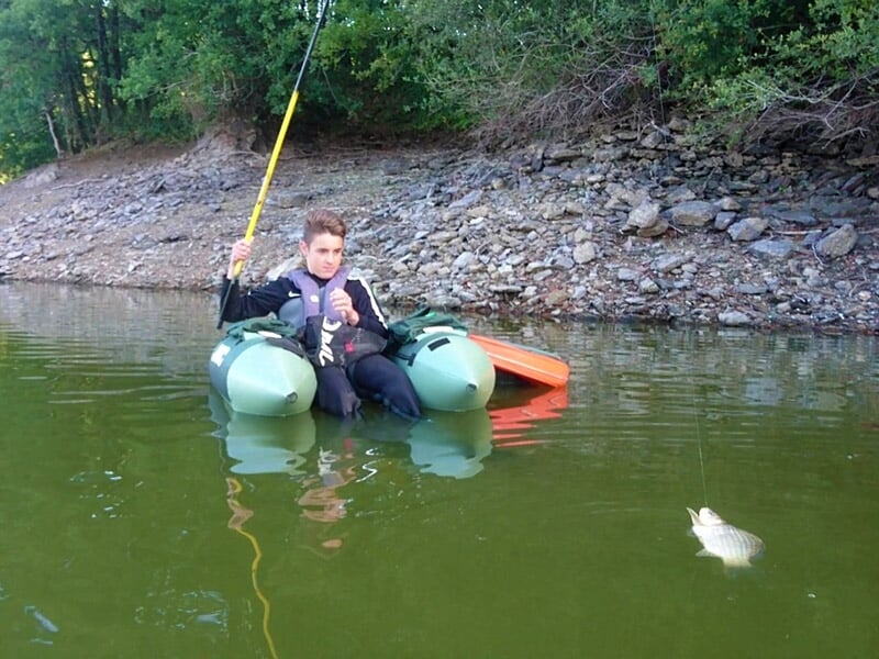 Jeune garçon assis sur un float tube dans un lac, tenant une canne à pêche avec un poisson dans l'eau.