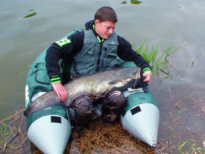 Adolescent assis sur un float tube dans un plan d'eau, tenant un grand poisson qu'il vient de pêcher.