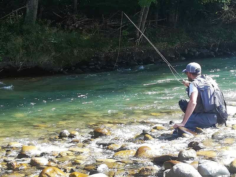 Homme assis au bord d'une rivière claire, pêchant à la canne dans un environnement naturel paisible et boisé.