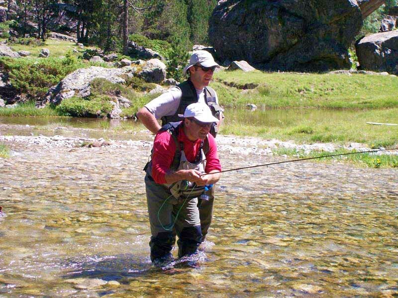 Deux pêcheurs en waders dans une rivière peu profonde, l'un enseignant la pêche à l'autre sous un ciel ensoleillé.