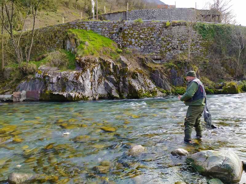 Pêcheur debout dans une rivière avec un mur en pierre en arrière-plan, lançant sa ligne dans l'eau claire.