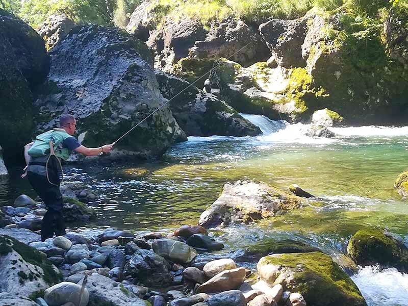 Pêcheur en waders lançant sa ligne dans une rivière rocheuse entourée de gros rochers et de végétation verte.