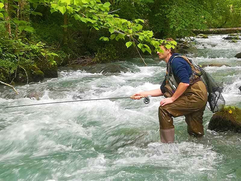 Pêcheur concentré dans une rivière agitée, tenant sa canne à pêche dans un cadre naturel verdoyant.