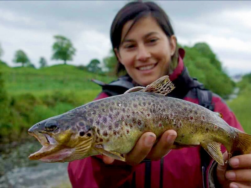 Femme souriante tenant une truite brune fraîchement pêchée, avec un paysage flou en arrière-plan.